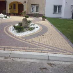 Okrągły podjazd z kostki brukowej w dwóch odcieniach, otaczający rabatę z białych kamieni i roślin, na tle dwóch budynków z numerami 10 i 11.