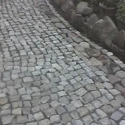 Ścieżka wyłożona drobną, szarą kostką. Po prawej stronie widoczny kamienny murek i zieleń.