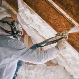 Pracownik w kombinezonie ochronnym aplikuje piankę izolacyjną na poddaszu, widoczne drewniane elementy konstrukcji dachu i świeżo nałożona, biała pianka. Dysza pistoletu natryskowego rozpyla pianę...