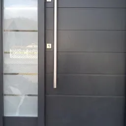 Ciemnoszare drzwi wejściowe z długim, pionowym uchwytem ze stali nierdzewnej i szklanym panelem bocznym, widoczne z bliska.