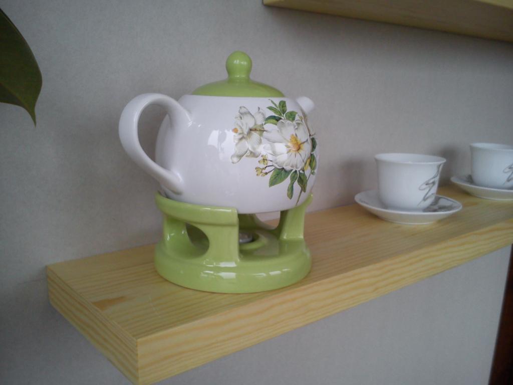Jasna, drewniana półka z czajnikiem na podgrzewaczu i filiżankami. Czajnik biały, zdobiony kwiatami, z zieloną pokrywką i podstawką. Minimalistyczny wystrój.