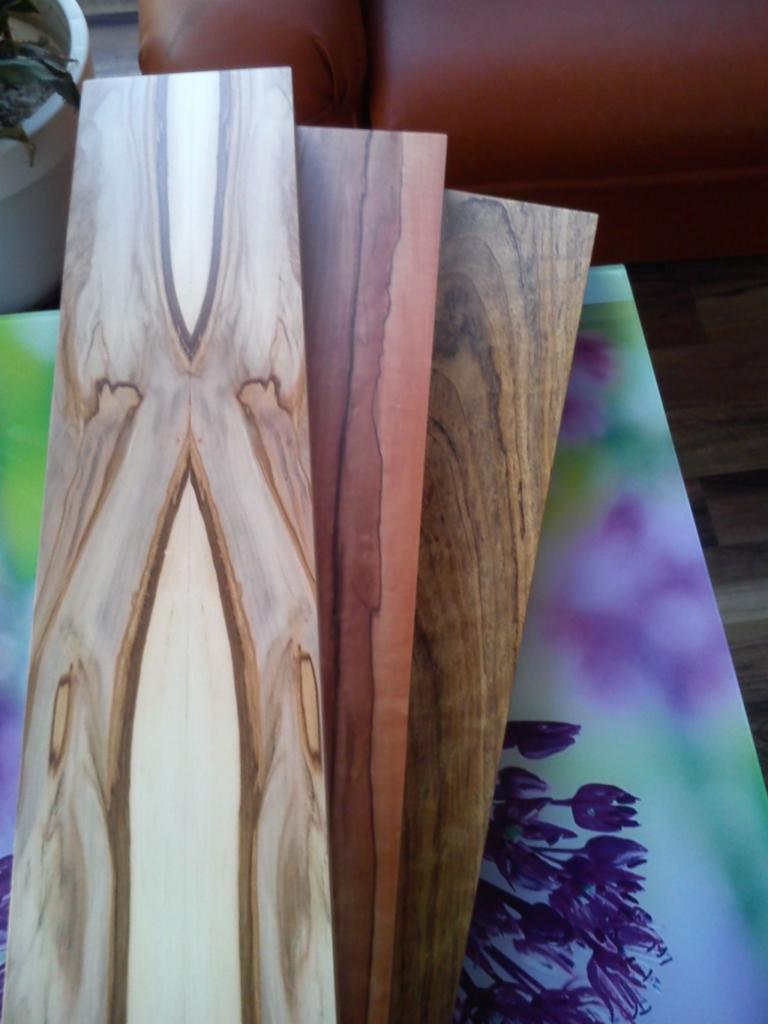 Trzy próbki tarcicy o różnej fakturze i kolorystyce drewna, ułożone na blacie ze wzorem kwiatowym. Widoczny fragment skórzanej sofy w tle.