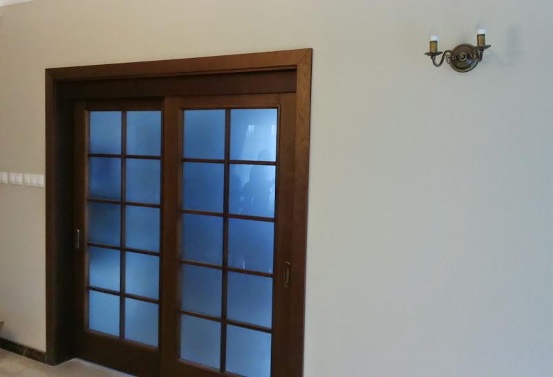 Drewniane drzwi przesuwne z szybami w kratkę, oświetlone przez kinkiet na ścianie. Widoczne detale wykończenia i naturalne światło wpadające przez szyby.