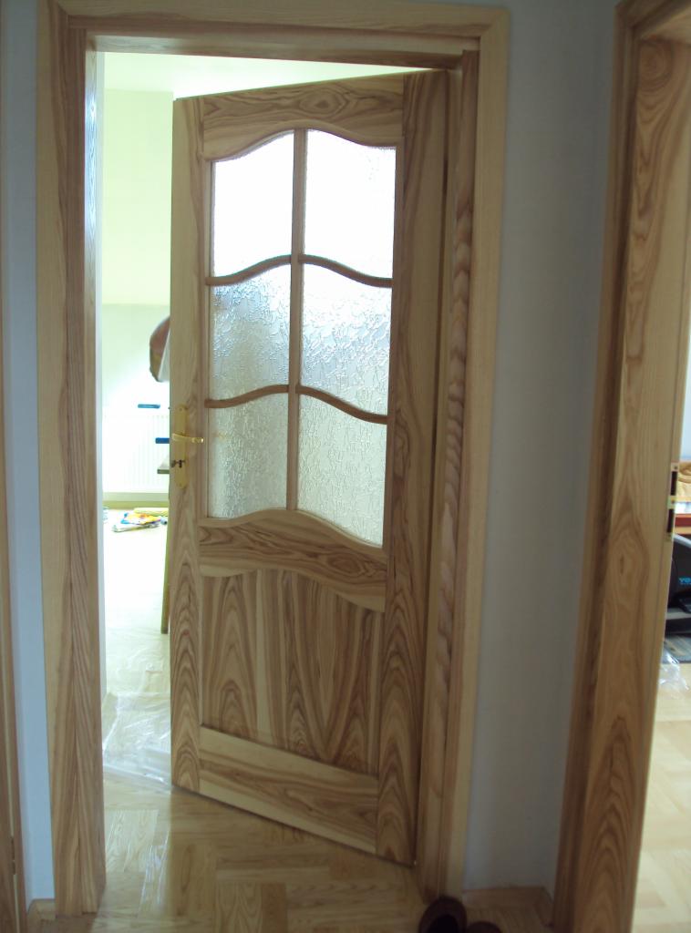 Otwarta, drewniana drzwi wewnętrzne z szybą o falistym wzorze, widoczne z bliska, z jasnym wykończeniem i złotą klamką, na tle jasnego wnętrza.