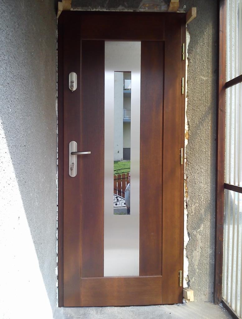 Brązowe drzwi zewnętrzne z pionowym, srebrnym panelem i widocznym zamkiem, zamontowane w surowym otworze ściany. Widoczny fragment elewacji i okna.