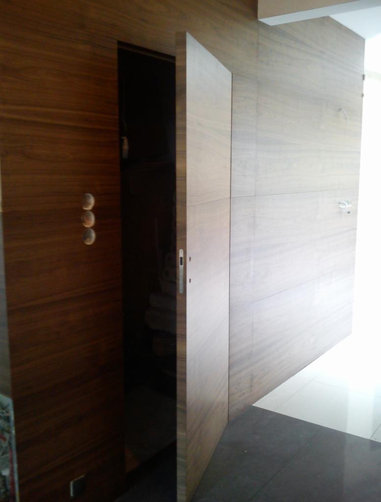 Otwarta, drewniana, wewnętrzna drzwi z trzema okrągłymi uchwytami na tle ściany z drewnianą okładziną i jasnej podłogi.