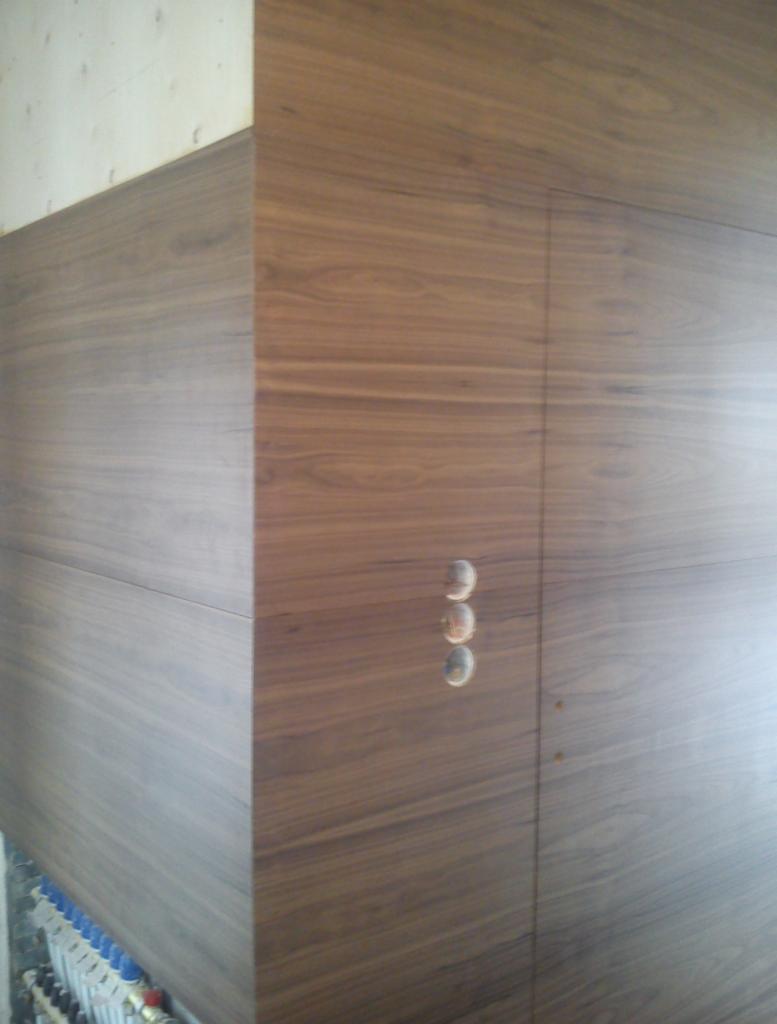 Ukryte drzwi z forniru orzechowego z widocznymi otworami na klamkę, integrujące się ze ścianą. Detal wykończenia wnętrza.