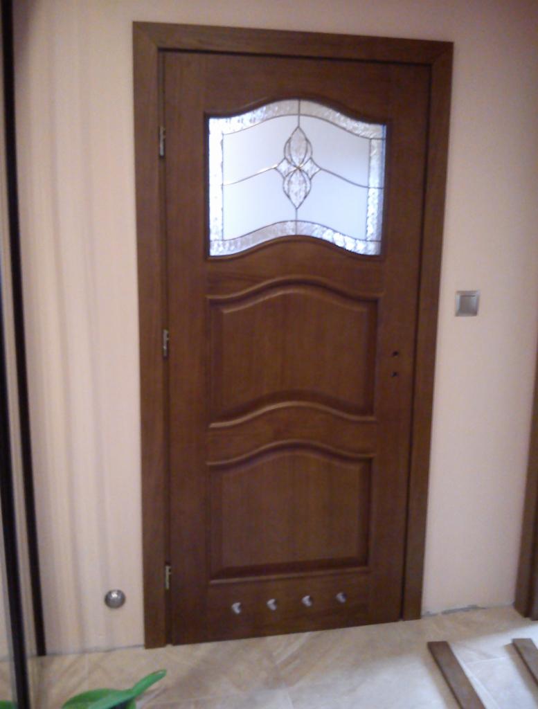 Drewniane drzwi wewnętrzne z ozdobnym szkłem i delikatnymi frezami, zamontowane w jasnym wnętrzu, widoczne małe haczyki na dole drzwi.