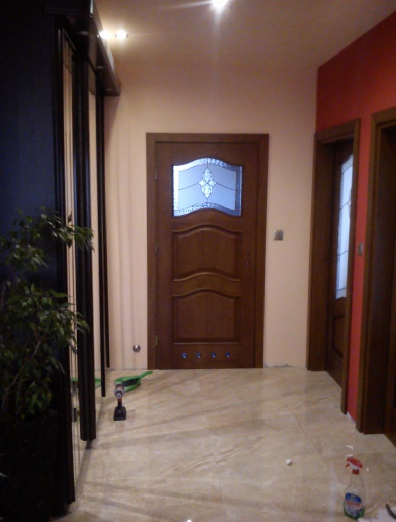 Montaż brązowych drzwi wewnętrznych z szybą dekoracyjną w przedpokoju z beżową podłogą. Widoczna wkrętarka i butelka z płynem do czyszczenia.