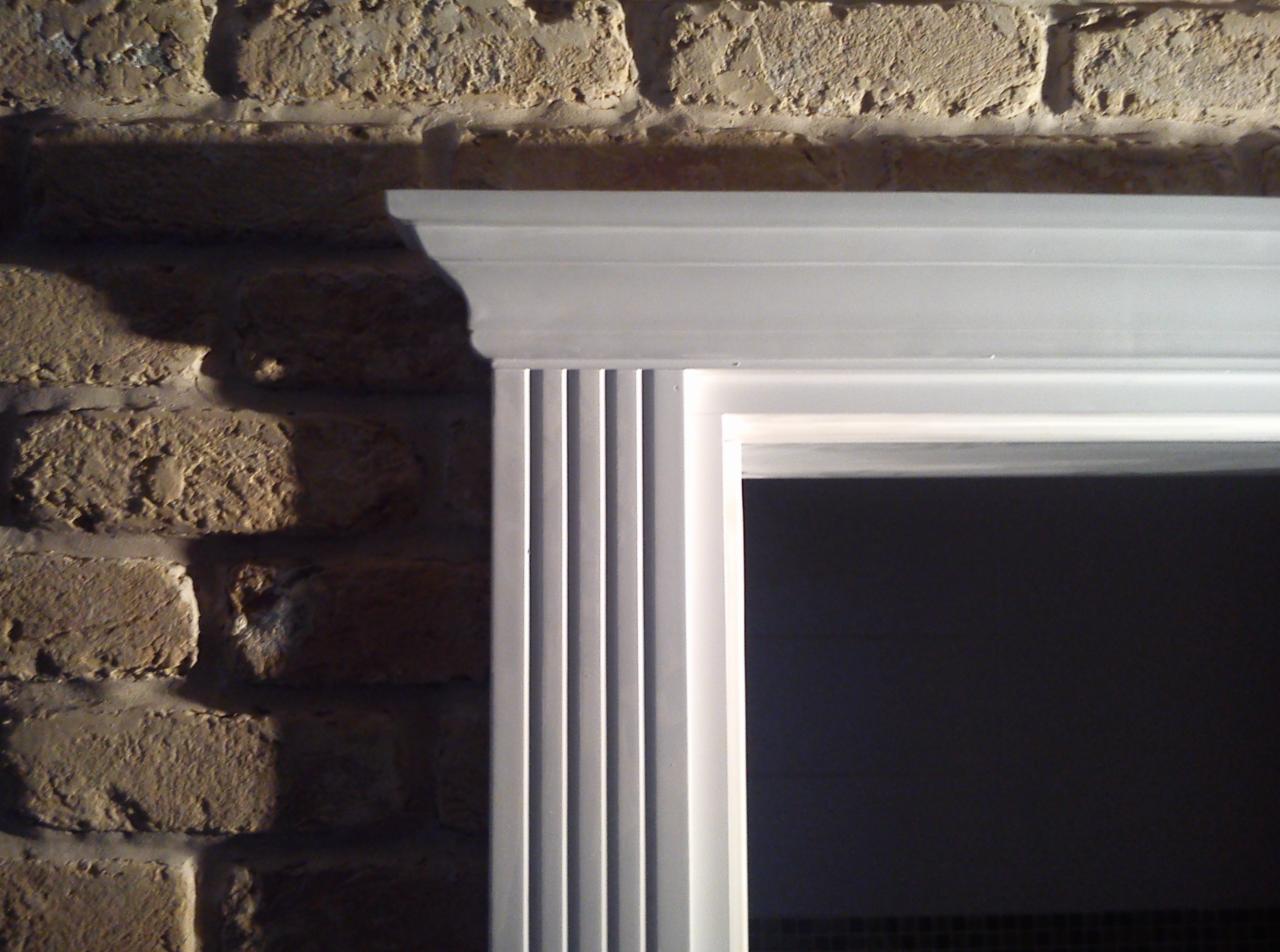 Biała rama drzwi z dekoracyjnymi żłobieniami, przylegająca do ściany z cegły. Widoczne detale wykończenia i połączenie materiałów.