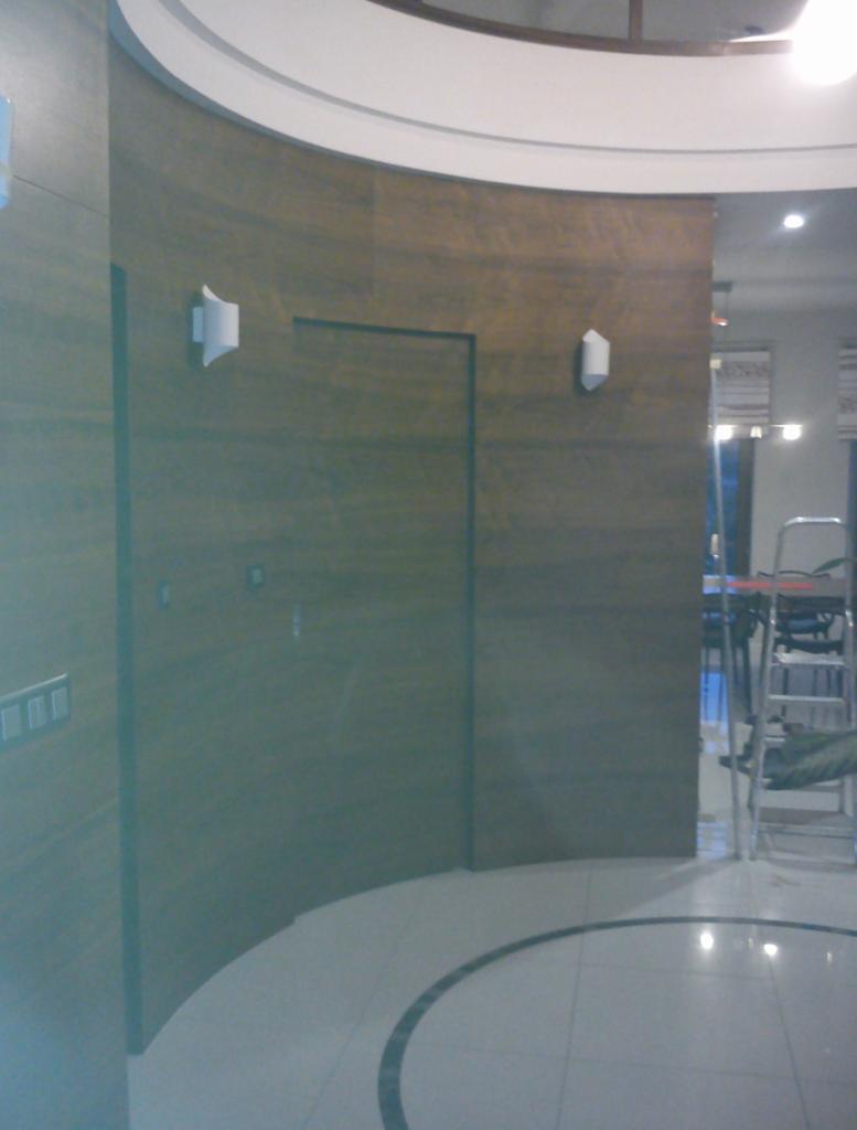 Zakrzywiona ściana w odcieniach brązu z ukrytymi drzwiami i dwoma kinkietami. Podłoga wyłożona białymi płytkami z czarnym akcentem.