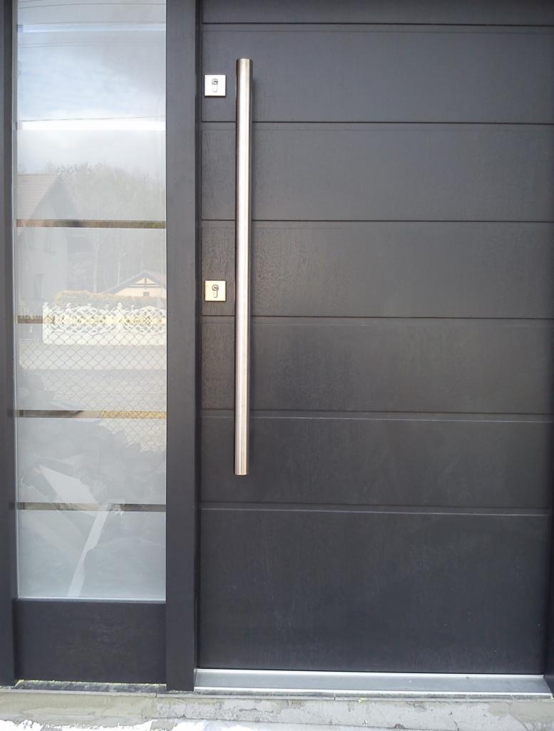 Ciemnoszare drzwi wejściowe z długim, pionowym uchwytem ze stali nierdzewnej i szklanym panelem bocznym, widoczne z bliska.