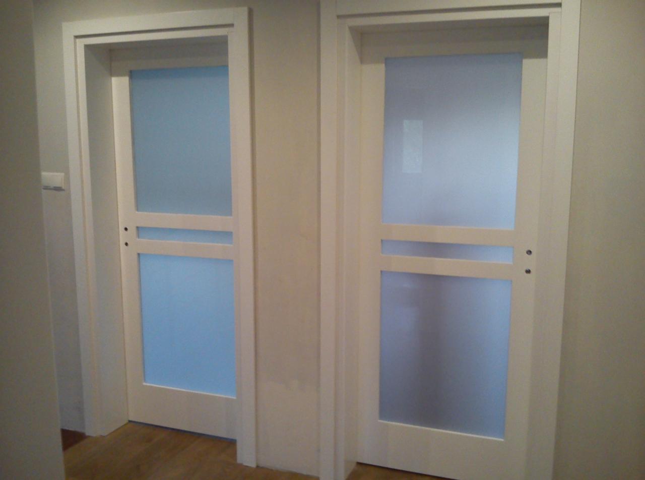 Dwa białe, wewnętrzne drzwi z matowym szkłem i poziomymi listwami, zamontowane w jasnym pomieszczeniu z drewnianą podłogą. Widok na korytarz.