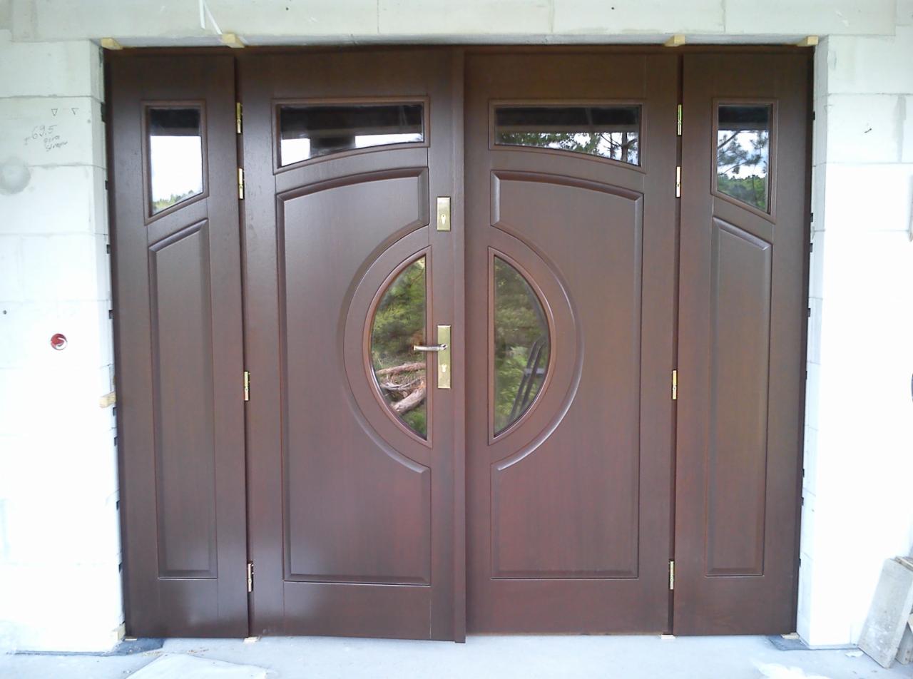 Brązowe, dwuskrzydłowe drzwi wejściowe z okrągłymi szybami, zamontowane w surowym murze. Widoczne zawiasy i klamka.