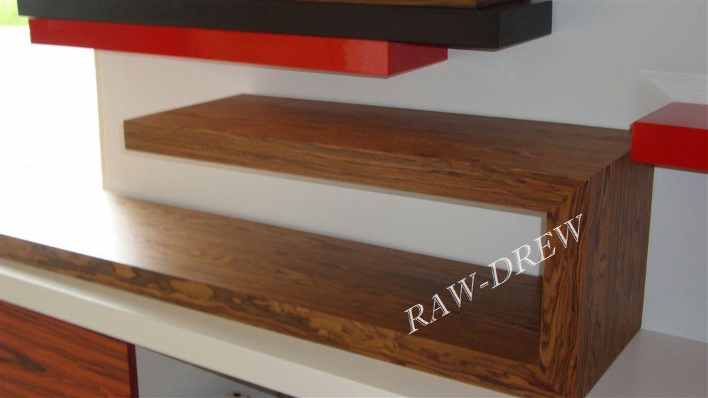 Drewniane półki w nowoczesnym stylu, z wyraźnym usłojeniem drewna, wkomponowane w białą zabudowę, z elementami w kolorze czarnym i czerwonym. Nietypowe ujęcie.