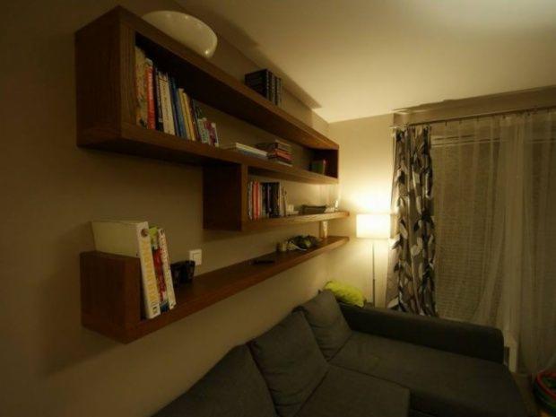 Drewniane półki ścienne z książkami w salonie, oświetlone lampą podłogową. Widoczna szara sofa i zasłony w tle. Ciepłe oświetlenie wnętrza.