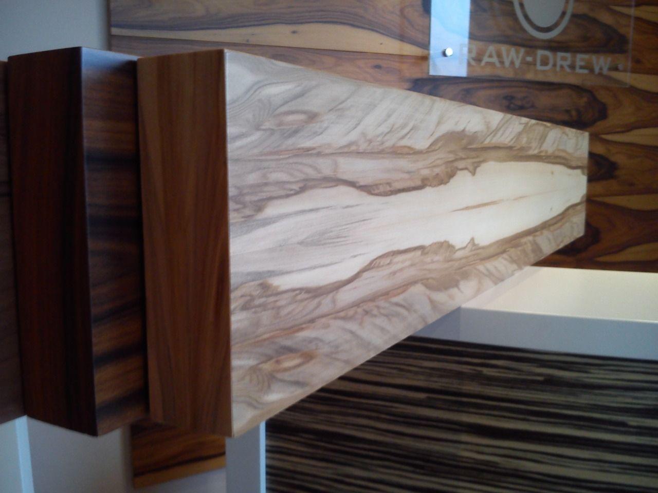 Drewniane próbki drzwi o różnym wykończeniu i usłojeniu, prezentowane pod kątem na tle ściany z drewnianą okładziną i logo firmy Raw-Drew.