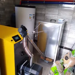 Żółto-szary piec na pellet z podajnikiem i zasobnikiem, obok worki pelletu, rury CO na ścianie z płytek.