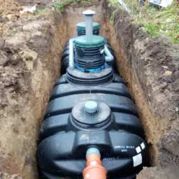 Czarny, żebrowany zbiornik na szambo lub deszcz&oacute;wkę, częściowo zakopany w wąskim wykopie z widocznymi rurami przyłączeniowymi i etykietami informacyjnymi na boku, z ziemią i roślinnością w tle.