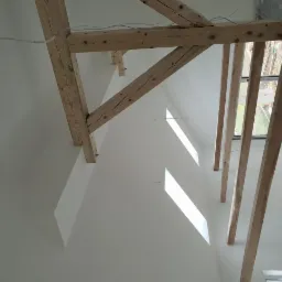 Białe, skośne poddasze z drewnianą konstrukcją stropu i belkami, widok z dołu, okno dachowe w tle.