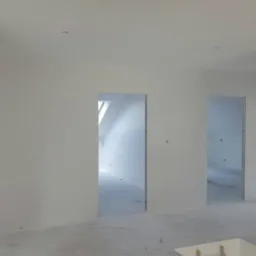 Wnętrze w trakcie prac wykończeniowych, świeżo wyszpachlowane ściany w odcieniach bieli, widoczne surowe otwory drzwiowe i okienne, przygotowane do dalszych prac malarskich i montażu stolarki.