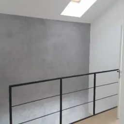 Wnętrze poddasza z oknem dachowym, szarą ścianą z efektem betonu i czarną, minimalistyczną balustradą.