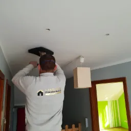 Pracownik w szarej bluzie z logo firmy montuje czarną oprawę oświetleniową w białym suficie, widoczne otwory na inne lampy, wnętrze w trakcie remontu z zielonym pokojem w tle.