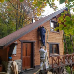 Dwaj mężczyźni na drabinie ocieplają drewniany dom z zewnątrz, używając węża do wdmuchiwania izolacji, widoczne drewniane deski i narzędzia na podeście.