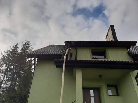 Pracownik w kombinezonie wdmuchuje izolację celulozową w poddasze domu jednorodzinnego przez długą rurę z poziomu dachu, widoczny fragment elewacji w kolorze pistacjowym.