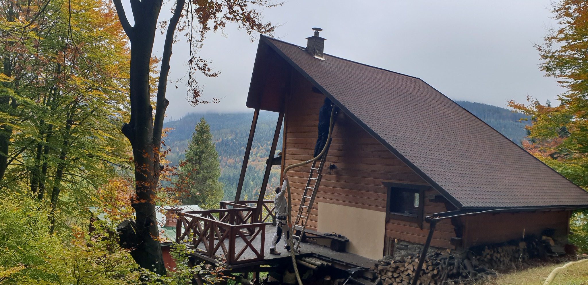 Pracownicy ocieplają drewniany dom z zewnątrz, używając rury do wdmuchiwania izolacji, widok na góry w tle.