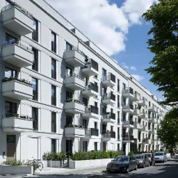 Nowoczesny budynek mieszkalny z białymi balkonami, widok z perspektywy ulicy, z zaparkowanymi samochodami i rowerem przed wejściem.