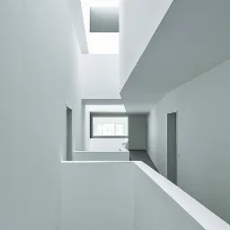 Wnętrze budynku z białymi ścianami i stropami, widok z góry na korytarz z oknem w tle, prosta architektura.