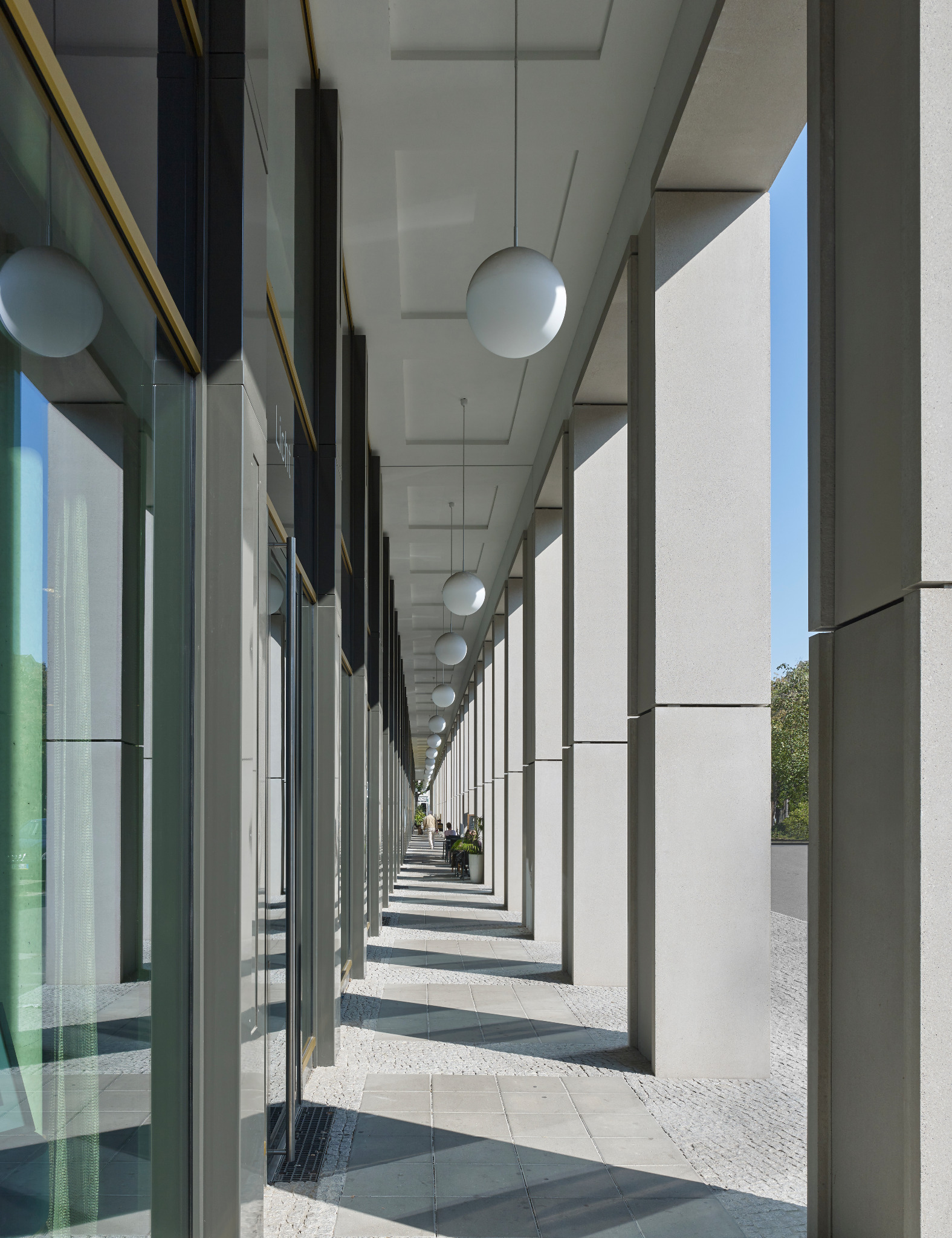 Długi pasaż handlowy z rzędem kolumn i wiszących kulistych lamp, widok w perspektywie z odbiciami w szklanych witrynach.