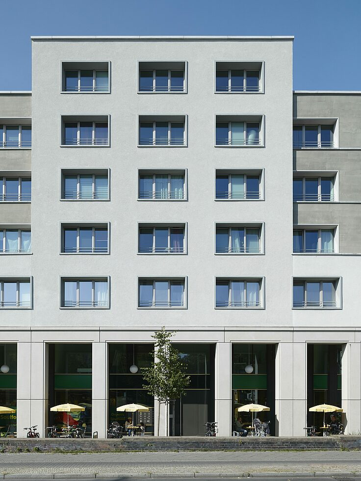 Fasada nowoczesnego, wielopiętrowego budynku usługowego z licznymi oknami i lokalami handlowymi na parterze z parasolami i stolikami na zewnątrz, widoczna mała roślina na tle wejścia.