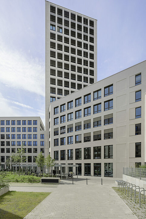 Nowoczesny kompleks biurowy z wysokim wieżowcem i niższymi budynkami, widok z dziedzińca z kostką brukową i zielenią, okna z roletami.