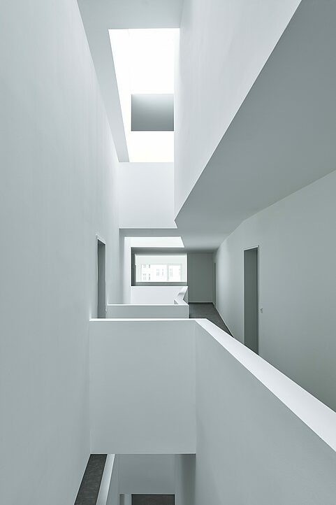 Wnętrze budynku z białymi ścianami i stropami, widok z góry na korytarz z oknem w tle, prosta architektura.