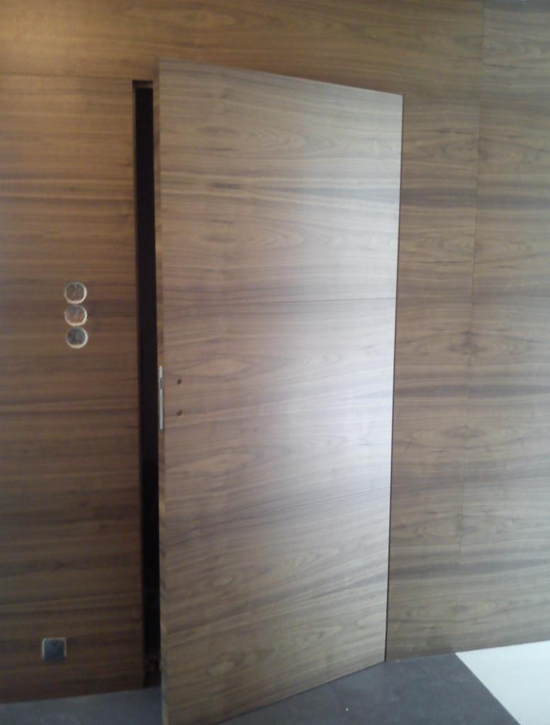 Drewniana szafa wnękowa z przesuwnymi drzwiami, zlicowana ze ścianą o podobnym wzorze drewna, widoczne detale wykończenia i minimalistyczny uchwyt.