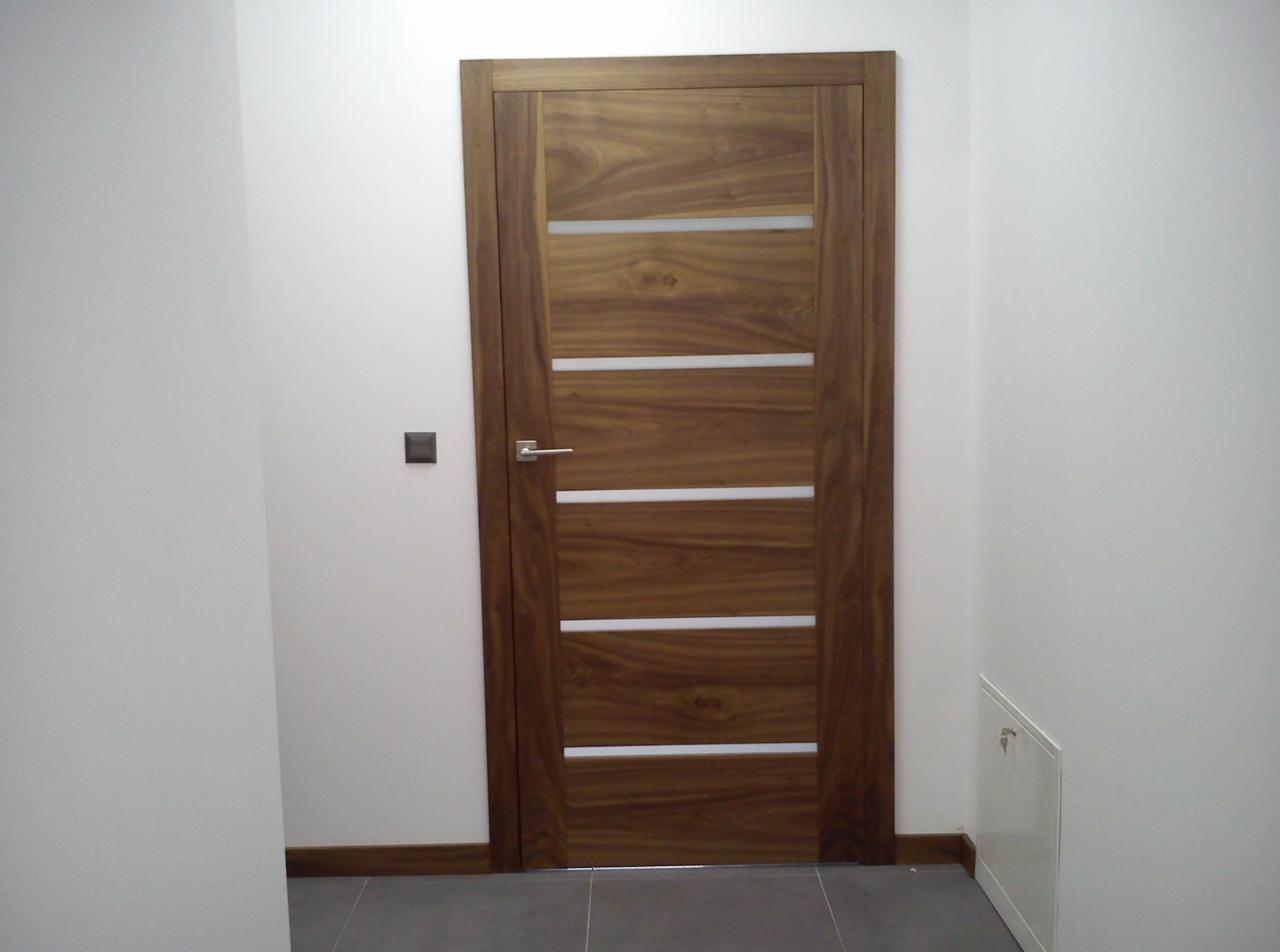 Drewniane drzwi wewnętrzne z poziomymi, mlecznymi przeszkleniami, osadzone w białej ścianie z szarą podłogą, widoczny włącznik światła i mała szafka rewizyjna.