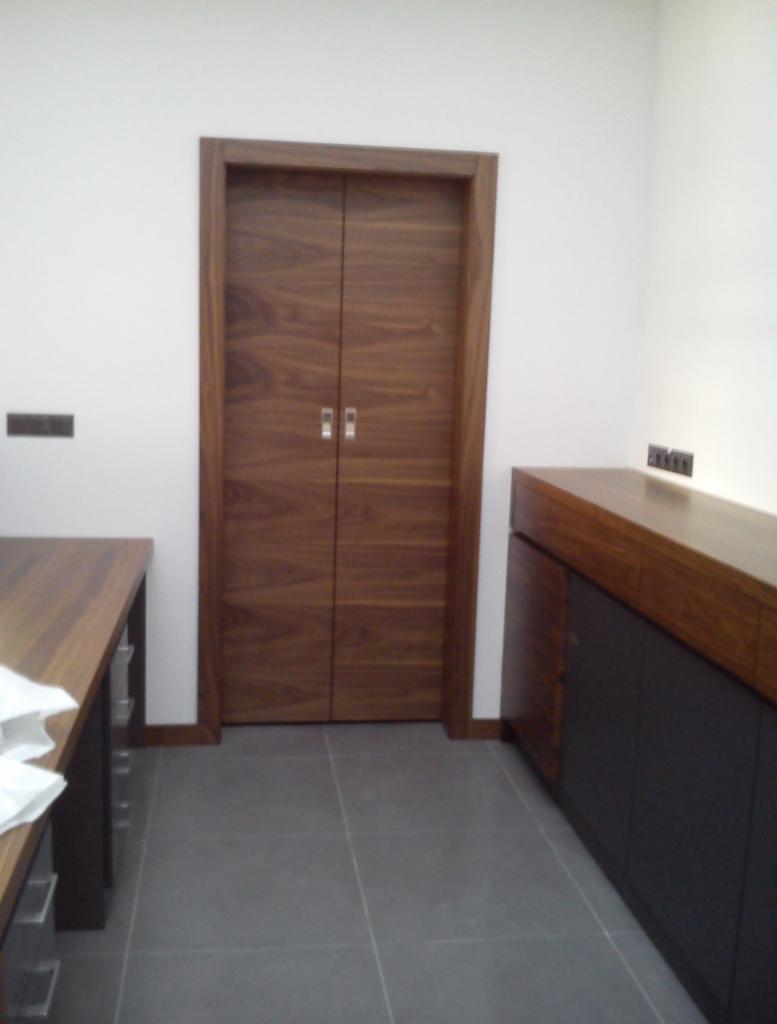 Dwuskrzydłowe drzwi z ciemnego drewna z prostokątną ramą, dopasowane kolorystycznie do blatu komody i biurka z szufladami w minimalistycznym wnętrzu z szarą podłogą.