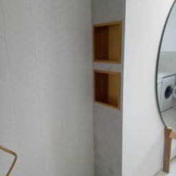 KMR-BUD Mariusz Moskal - Nowoczesna kabina prysznicowa z białymi płytkami ułożonymi w jodełkę, złotą armaturą i wnękami w ścianie wykończonymi drewnem, odbicie pralki w okrągłym lustrze.