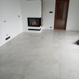 KMR-BUD Mariusz Moskal - Wykończony salon z szarą, matową podłogą z płytek, nowoczesnym kominkiem z czarnymi akcentami i ułożonym drewnem, oraz drewnianymi drzwiami w tle.