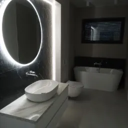 Nowoczesna łazienka w odcieniach szarości z okrągłym lustrem podświetlonym LED, owalną umywalką nablatową na szafce z marmurowym blatem, wolnostojącą wanną i toaletą.