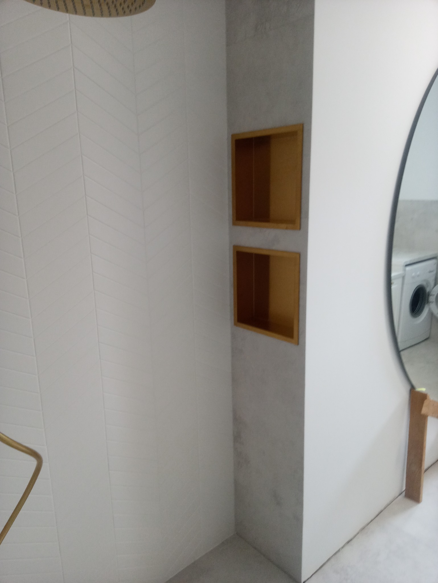 Nowoczesna kabina prysznicowa z białymi płytkami ułożonymi w jodełkę, złotą armaturą i wnękami w ścianie wykończonymi drewnem, odbicie pralki w okrągłym lustrze.