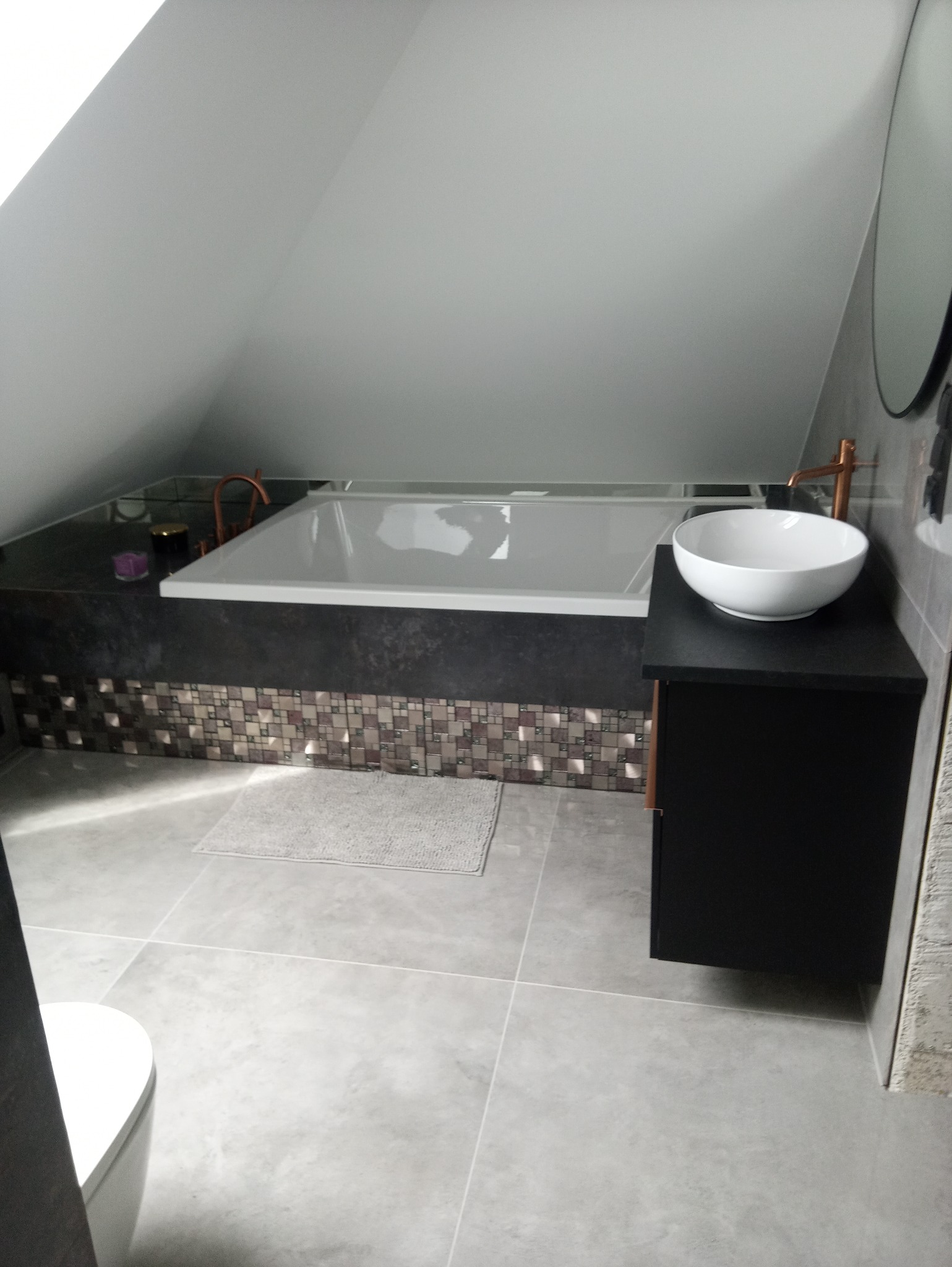 Nowoczesna łazienka na poddaszu z białą wanną obudowaną mozaiką, umywalką nablatową na czarnej szafce i baterią w kolorze miedzi, szare płytki podłogowe i częściowo widoczna toaleta.