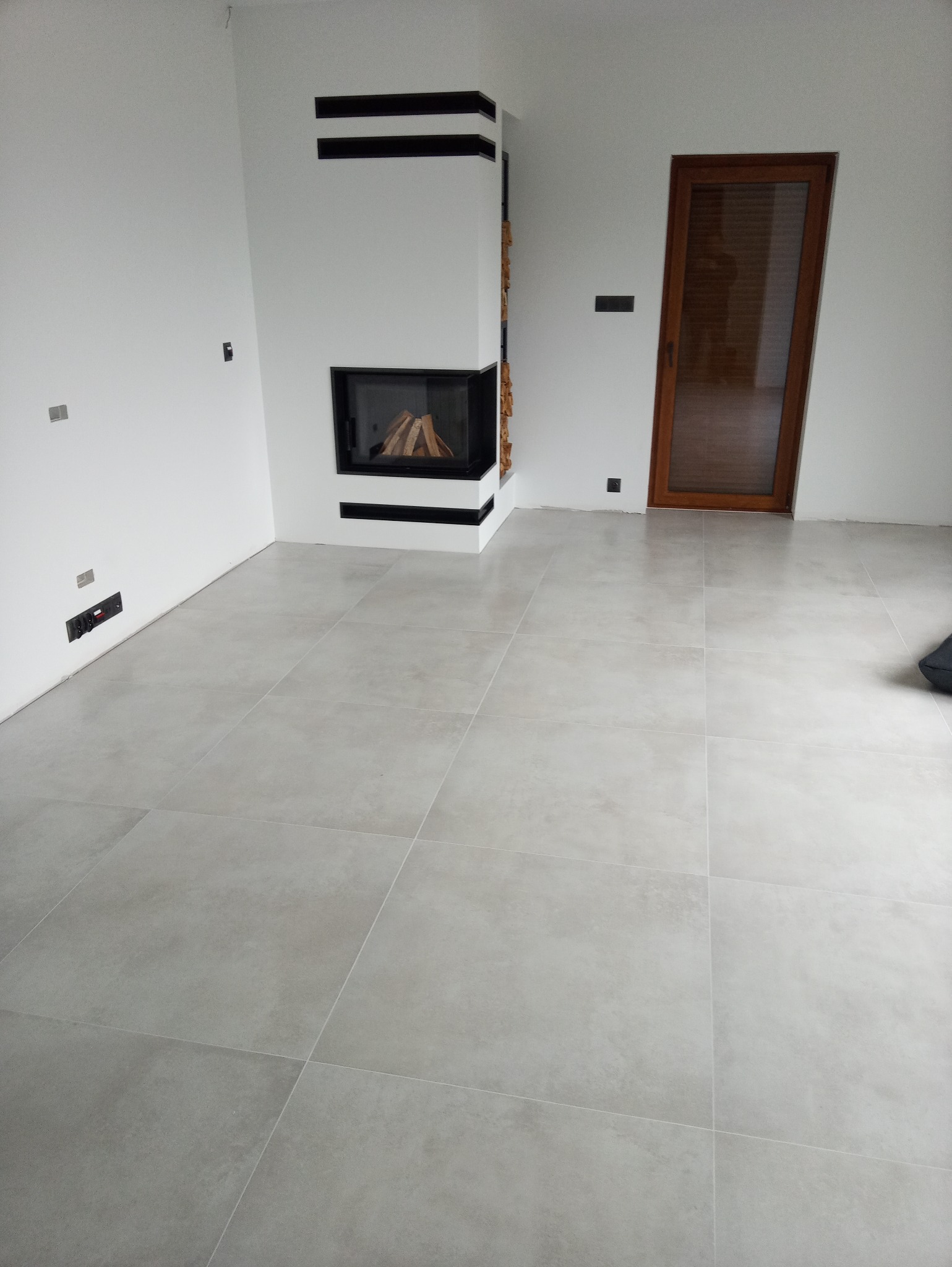 Wykończony salon z szarą, matową podłogą z płytek, nowoczesnym kominkiem z czarnymi akcentami i ułożonym drewnem, oraz drewnianymi drzwiami w tle.