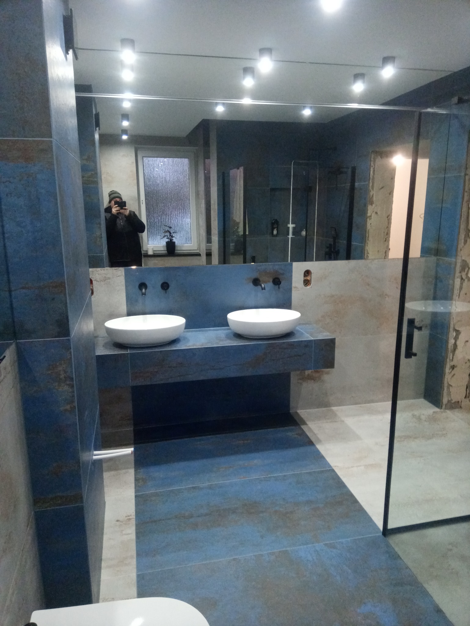 Nowoczesna łazienka z dwoma białymi umywalkami nablatowymi na niebieskim blacie imitującym kamień, duże lustro z odbiciem osoby robiącej zdjęcie telefonem, kabina prysznicowa ze szklaną ścianką...