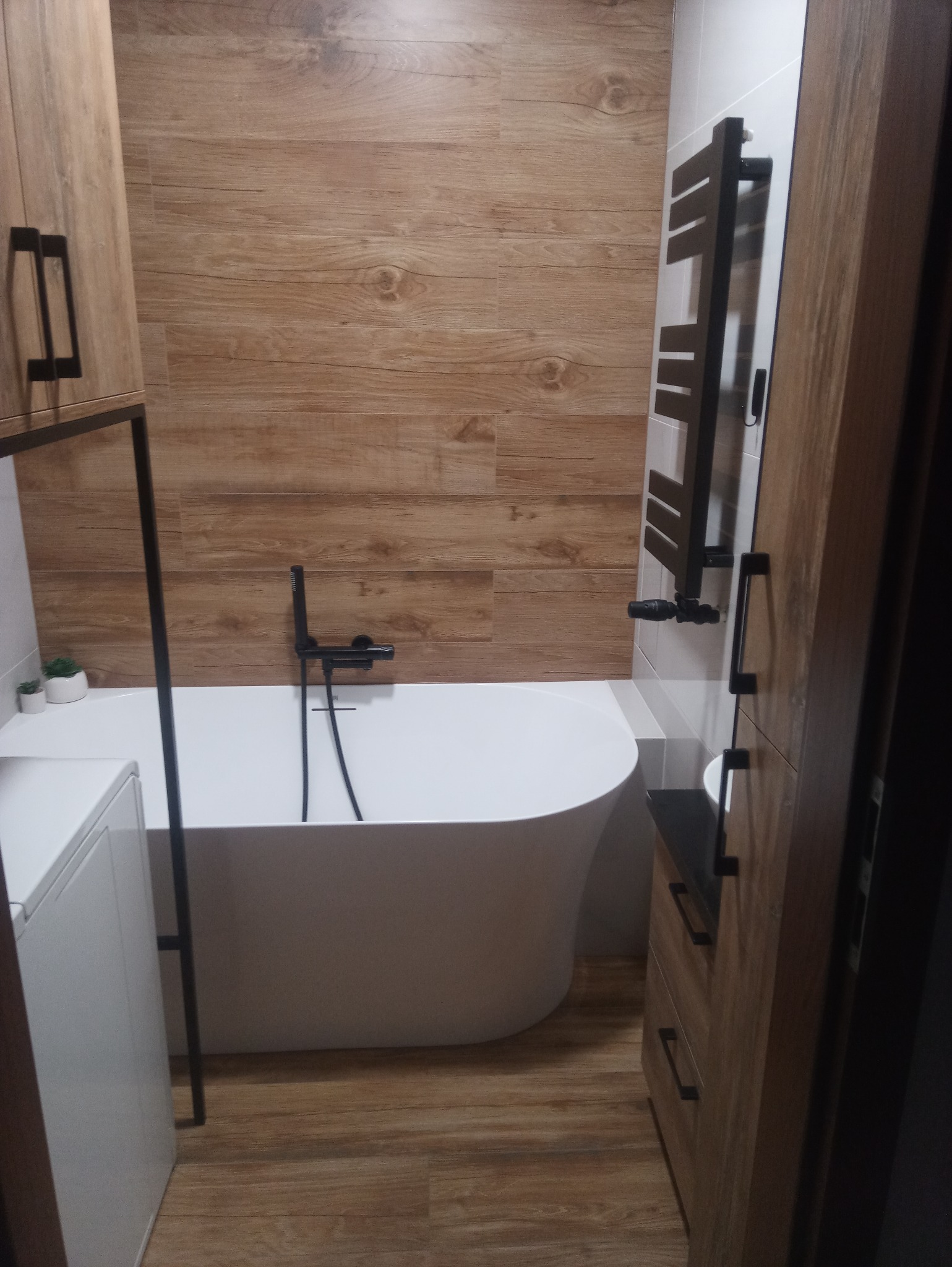 Nowoczesna łazienka z wolnostojącą białą wanną, czarną armaturą i grzejnikiem drabinkowym na tle drewnopodobnych płytek ściennych i podłogowych, uzupełniona szafką z blatem.