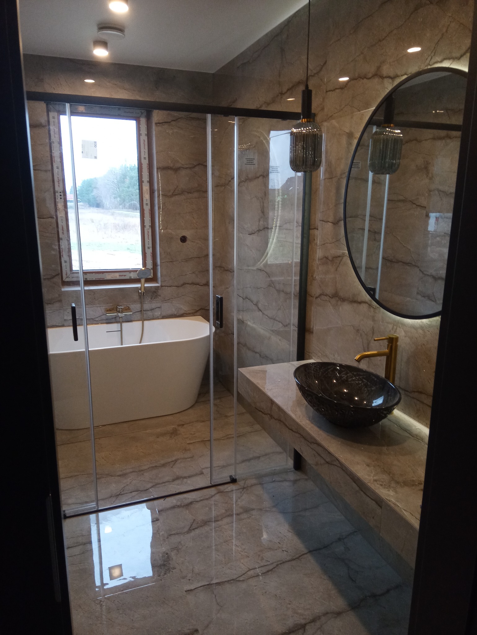 Nowoczesna łazienka z marmurowymi płytkami, owalną wanną przy oknie, szklaną kabiną prysznicową i umywalką w kształcie misy na blacie.