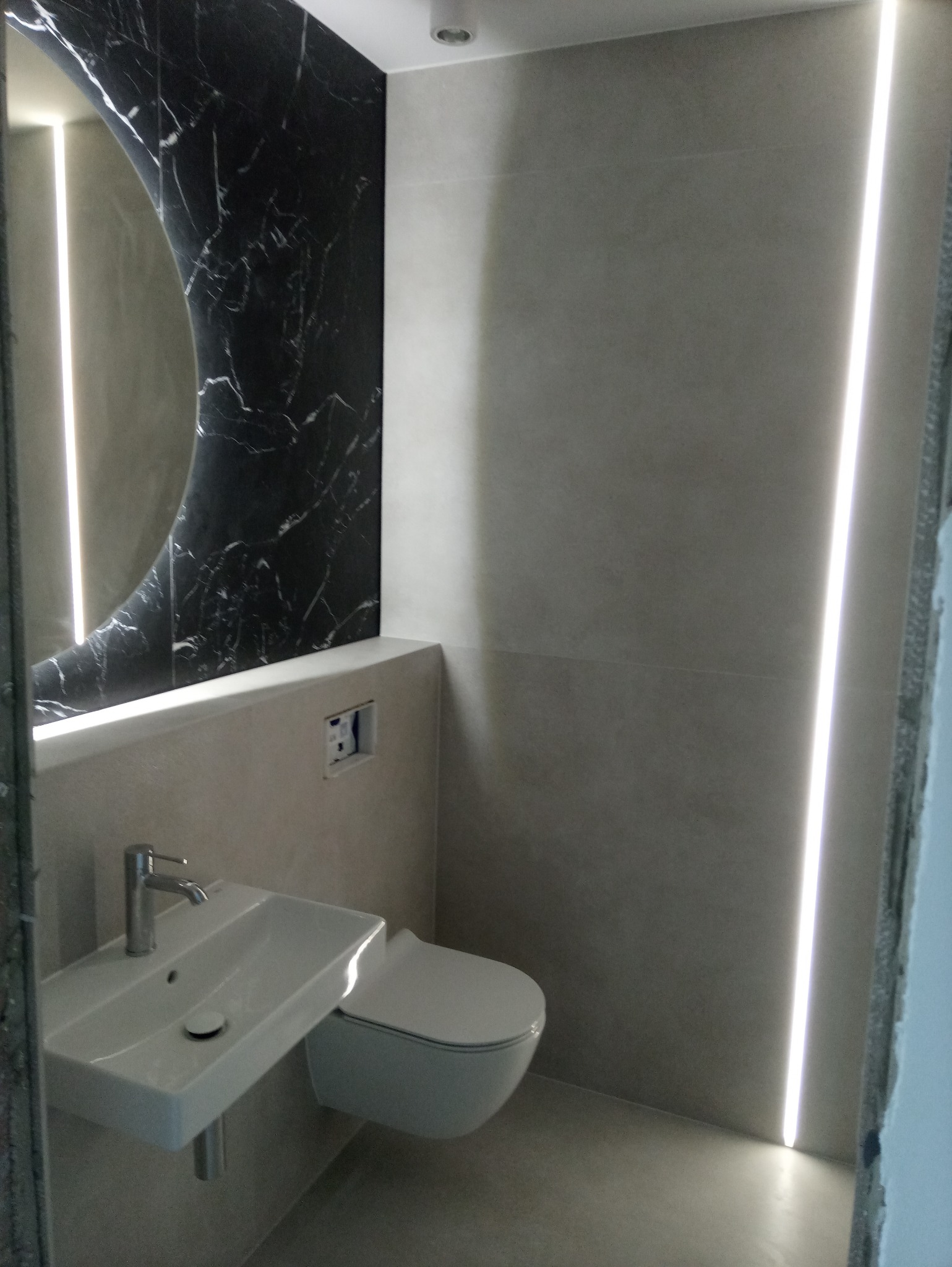 Nowoczesna łazienka z umywalką nablatową, podwieszaną toaletą i czarną ścianą z marmurowym wzorem, oświetlona listwą LED w pionowej szczelinie ściany.