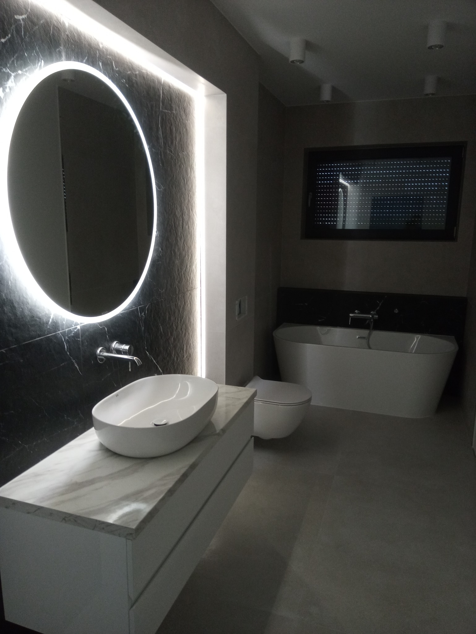 Nowoczesna łazienka w odcieniach szarości z okrągłym lustrem podświetlonym LED, owalną umywalką nablatową na szafce z marmurowym blatem, wolnostojącą wanną i toaletą.